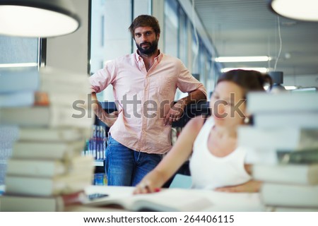 Asian university student girl sitting at the library desk with pile of books, international students preparing for exams while studying in library, brunette hair hindu man standing near bookshelves
