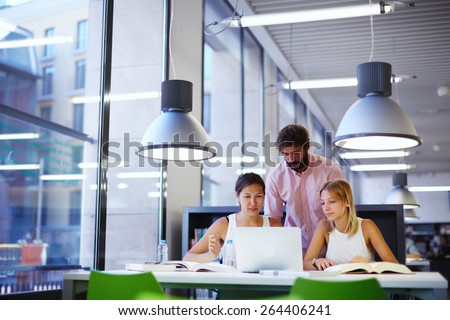 Group of international university students learning in library, three colleagues of modern work co-working space talking and smiling while sitting at the desk with laptop computer, exam preparation