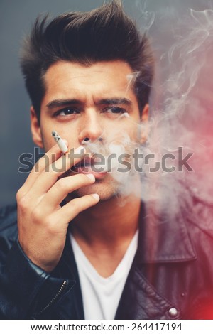 Close up portrait of handsome fashion man exhaling cigarette smoke while looking away, trendy attractive man blowing smoke out of his mouth standing on grey background, filtered image, red flare light