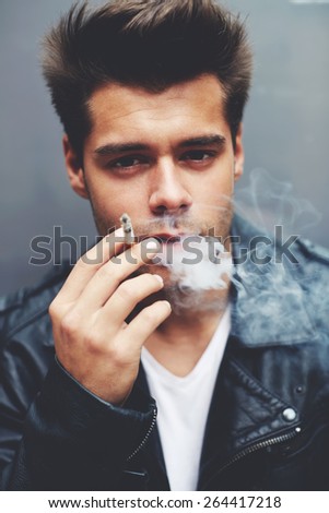 Close up portrait of handsome fashionable man exhaling cigarette smoke while looking to the camera, trendy attractive man blowing smoke out of his mouth standing on grey background, filtered image