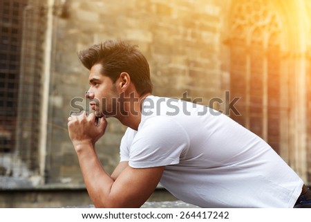 Side shot portrait of handsome man resting chin on clasped hands, confident man thinking about his choice, pensive man standing against gothic building, flare sun light, filtered image