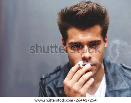 Close up portrait of handsome fashionable man exhaling cigarette smoke while looking to the camera, trendy attractive man blowing smoke out of his mouth standing on grey background, filtered image