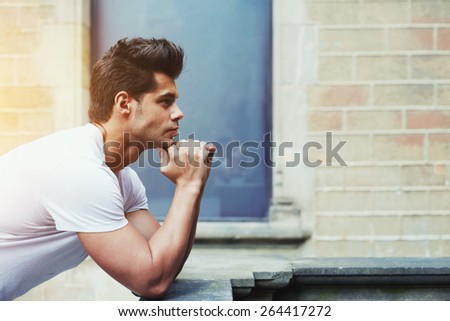 Side shot portrait of handsome man resting chin on clasped hands, confident man thinking about his choice, pensive man standing on brick wall background, flare sun light, filtered image