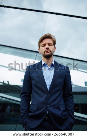 Half length of young businessman standing with closed eyes and hands in his pockets against office building, peaceful man having big hope looking confident