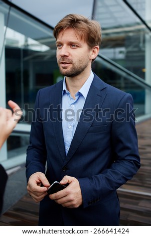 Portrait of handsome business man in suit talking to someone holding cellphone in the hands,young caucasian executive having conversation outside office during work break,colleagues having discussion