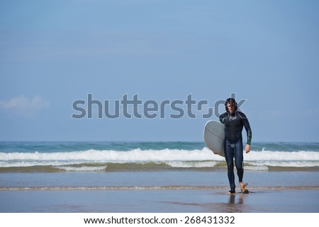 Young surfer sport man carrying his surfing board and going out the sea while on vacation during a sunny day, with an intense blue sky and big waves break on background