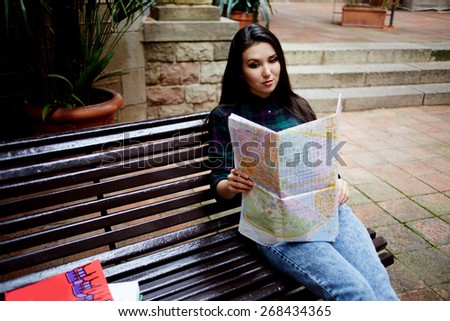Portrait of a young asian brunette hair woman holding a map while touring abroad, attractive tourist girl reading map and resting after walk in new city while sitting on the bench