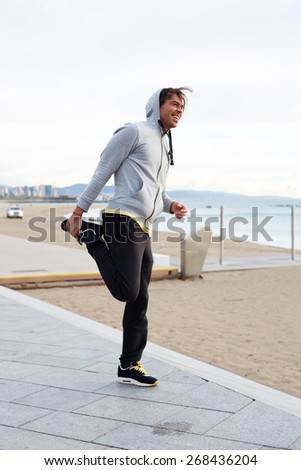 Full length shot of a young male jogger stretching before his run, male jogger doing stretching exercise while standing on seaside listening to music on headphones