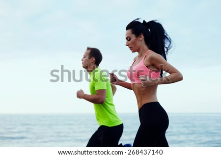 Side view of couple running outdoors, fit athlete man and fitness woman working out together running on beautiful beach with sea horizon on background