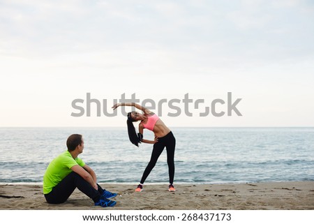 Young couple exercising and enjoying time outdoors, young fitness woman doing physical stretch exercise while her boyfriend taking break, couple working out on the beach at evening time