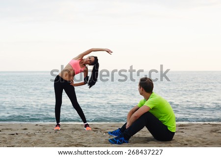 Young woman with beautiful figure doing stretching exercise on the beach, couple working out against the sea at evening time, athletic man taking a break while his girlfriend exercising