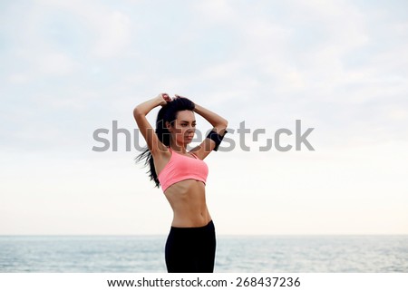 Portrait of attractive sports woman with beautiful figure getting ready for run, charming young woman having a rest during her fitness training on the beach, female runner standing on seashore resting