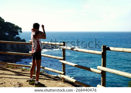 Side shot of handsome young runner stretching before starting his run while standing on edge of a cliff with a wooden fence and enjoying ocean view from altitude, cross process, filtered image