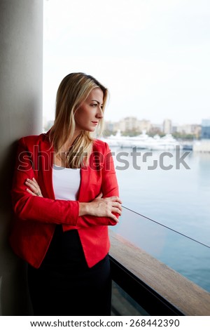 Portrait of pensive businesswoman looking out of an office balcony with beautiful seaport view on background, female executive with crossed arms thoughtfully looking away while having work break