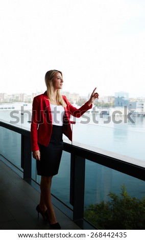 Elegant successful woman standing on the balcony of modern office building while calling for someone, rich well dressed woman standing in office with marina port view on background
