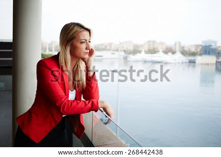 Portrait of pensive businesswoman looking out of an office balcony with beautiful seaport view on background, unhappy successful woman standing in exterior,blonde woman looking out of her window sadly
