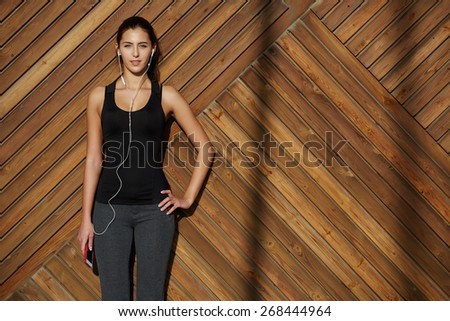 Attractive fit woman in sportswear listening to music with her headphones while standing on wooden background with space, young female runner enjoying the sun while listen to music though earphones