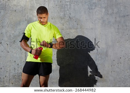 Dark-skinned male athlete in fluorescent t-shirt holding a water bottle looking tired and exhausted, sporty young man resting after workout standing on concrete wall background at sunny afternoon