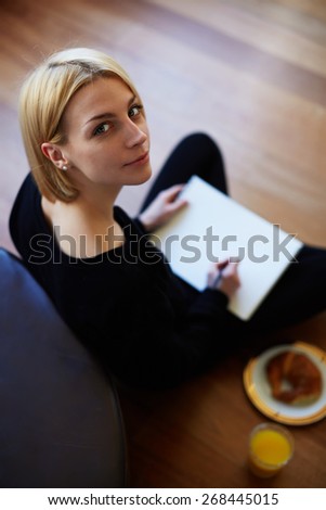 Top view shot of blonde hair young woman drawing on the paper while sitting on the floor of her living room, creative artist teenager girl having breakfast while painting on sheet of album paper