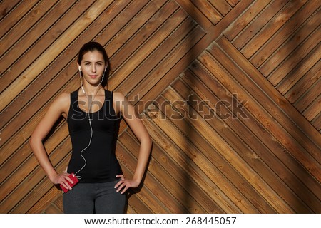 Attractive fit woman in sportswear listening to music with her headphones while standing on wooden background with space, young female runner enjoying the sun while listen to music though earphones