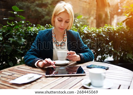 Young attractive businesswoman at coffee time working on digital tablet while sitting on the terrace of coffee shop, caucasian blond well dressed woman using touch pad while sitting in coffee shop