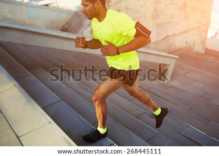 Cropped shot male dark-skinned athlete running up a flight of stairs with speed, sporty young man in fluorescent t-shirt training or working out outdoors while jogging up the steps, flare sun