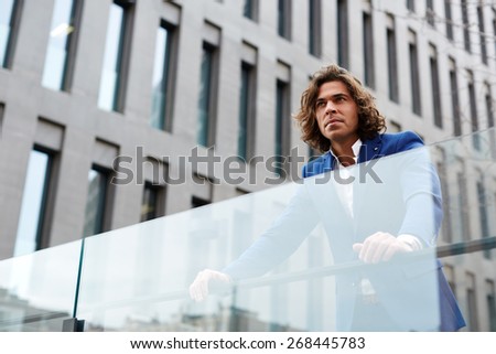 Portrait of handsome confident man leaning on glassy fence while standing against big office building in urban setting, attractive well dressed man looking away confident and concentrated