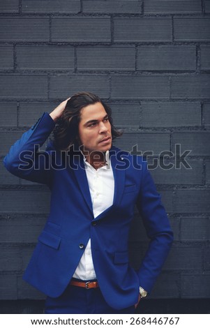 Portrait of long hair fashionable man in suit posing on black wall background, elegant and handsome adult man with hand above his head posing outdoors, filtered image