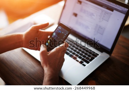 Close up male hands holding big smart phone while connecting to wireless, businessman using technology sitting at modern loft wooden desk, people and modern devices everywhere, flare sun light