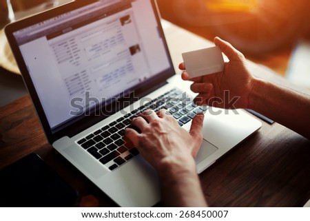 Rear view of male hands holding credit card typing numbers on computer keyboard while sitting at home at the wooden table, soft focus, flare sun light, cross process image