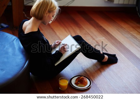 Beautiful young woman writing something in the note pad while sitting on the floor at living room, charming female studying doing homework at home, girl writes diary at her breakfast, flare light