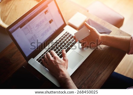 Rear view of male hands holding credit card typing numbers on computer keyboard while sitting at home at the wooden table, soft focus, flare sun light, cross process image