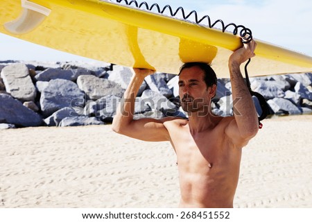 Portrait of handsome spanish brunette hair man with nude torso carrying big surfboard above his head while walking on the beach at beautiful sunny day, sexy men on the beach