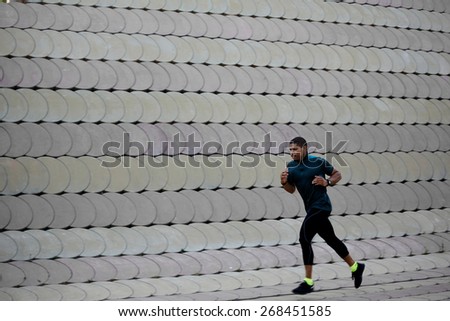 Full length portrait of athletic dark-skinned man running along beautiful grey wall outdoors, muscular build young runner working out while jogging in the park