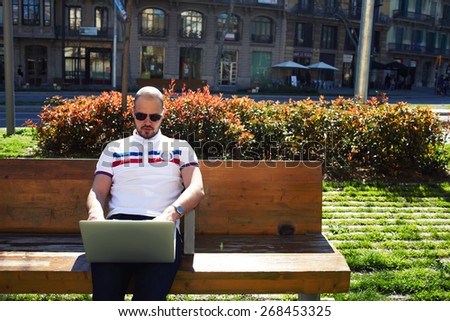 Young modern businessman freelancer working on his computer while sitting on city park bench, male student using notebook outdoors in urban setting while typing on keyboard, tourist working on laptop