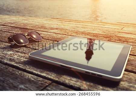 Sunglasses and digital tablet lying on wooden jetty at sunny day, touch pad wireless device, vacation travel and weekend break concept, flare sun light, focus on the sunglasses