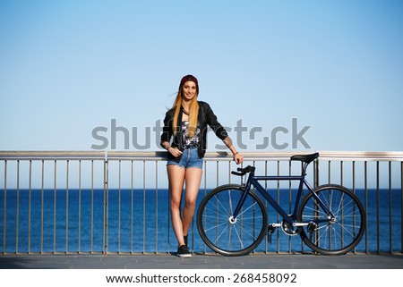 Full length portrait of stylish hipster girl standing with her sport fixed gear bike on the beach, young woman standing on the pier before ride along the seaside on her bicycle, promenade at sunny day