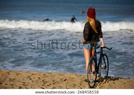 Attractive hipster girl relaxing on the beach after ride on her sport fixed gear bicycle, young woman standing on seashore looking to surfing people in water, charming female with rental bike outdoors