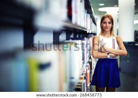 Portrait of a caucasian smiling female student holding book while standing in university library, pretty young woman standing near bookshelves in bookstore shop, exam preparation in university