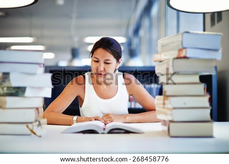 Entrained female student learning at the desk with a huge pile of study books in university library, young asian college student reading interesting textbook looking so absorbed with the content