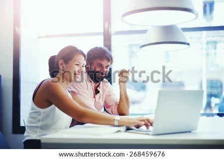 Couple of young designers working at modern office, two coworkers discussing fun project over a laptop, little team of businesspeople smiling and looking together at computer, students at library