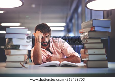Portrait of caucasian male student sitting at the desk behind stack of books looking exhausted and tired to study, teenager sitting at the table with a huge pile of study books in university library
