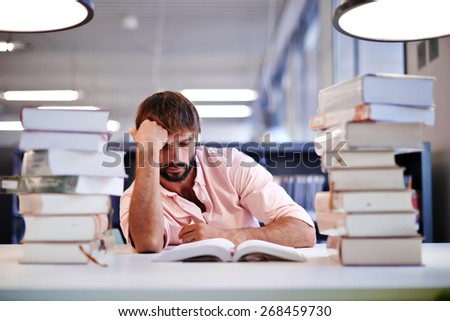 Frustrated male student sitting at the desk with a huge pile of study books in university library, young college student at hard exam preparation in study hall looking tired and weary