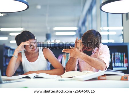 Two students having fun in library while studying at the desk with open books, brunette hair young man doing finger gun gesture to his asian classmate, hard preparation for university exams