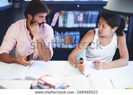 Portrait of young people studying together in the library, brunette asian female and spanish male students sitting at the desk with open books while prepare for exams in university study hall