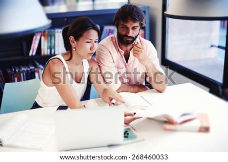 Two college students learning together at university library, brunette asian female and spanish male preparing for exams while sitting at the desk with open books and laptop computer in a study hall