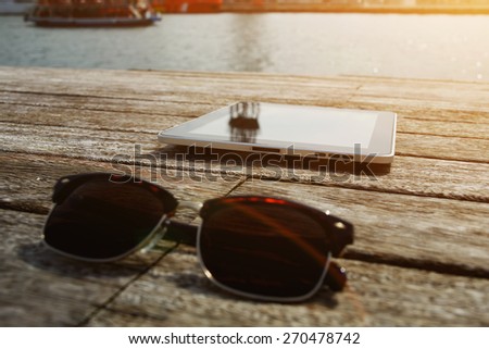 Sunglasses and digital tablet lying on wooden jetty with sailing tourist boat on background touch pad wireless device, vacation travel and weekend break concept