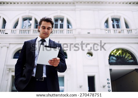 Confident man using digital tablet computer while standing in big light hall of railway station, successful businessman on way to work reading news on his touch pad, executive receiving news on email