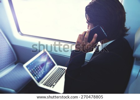 Businessman working busy with laptop on the way to work while sitting in train next to the window, handsome male having cell phone conversation, business man commuting to work while using technology