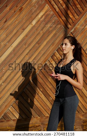 Attractive fit woman in sportswear listening to music with headphones while standing on wooden background with copy space, young female runner enjoying the sun while listen to music though earphones